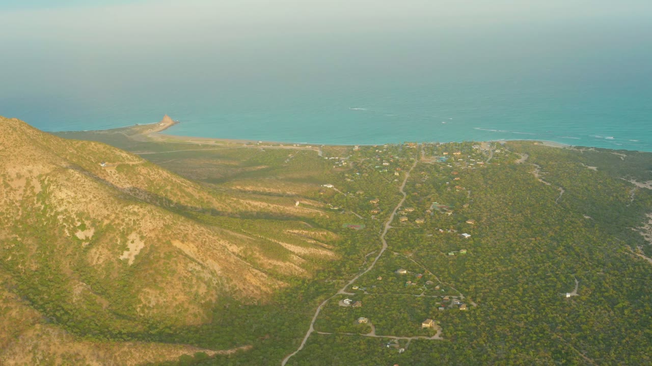 Cabo Pulmo aerial view with dry ridges on the left, darker green vegetation on the right, small buildings near the roads, a pale beach strip, blue ocean, and no visible people.
