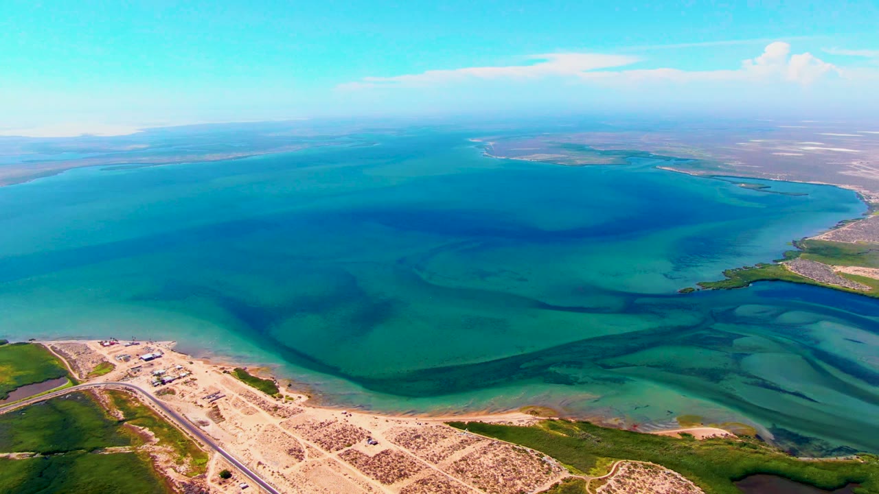 Aerial frame over San Carlos, Baja California Sur showing turquoise and deep blue coastal water, sandy shoreline, a road at lower left, low green vegetation, distant land, clouds, and no visible people.