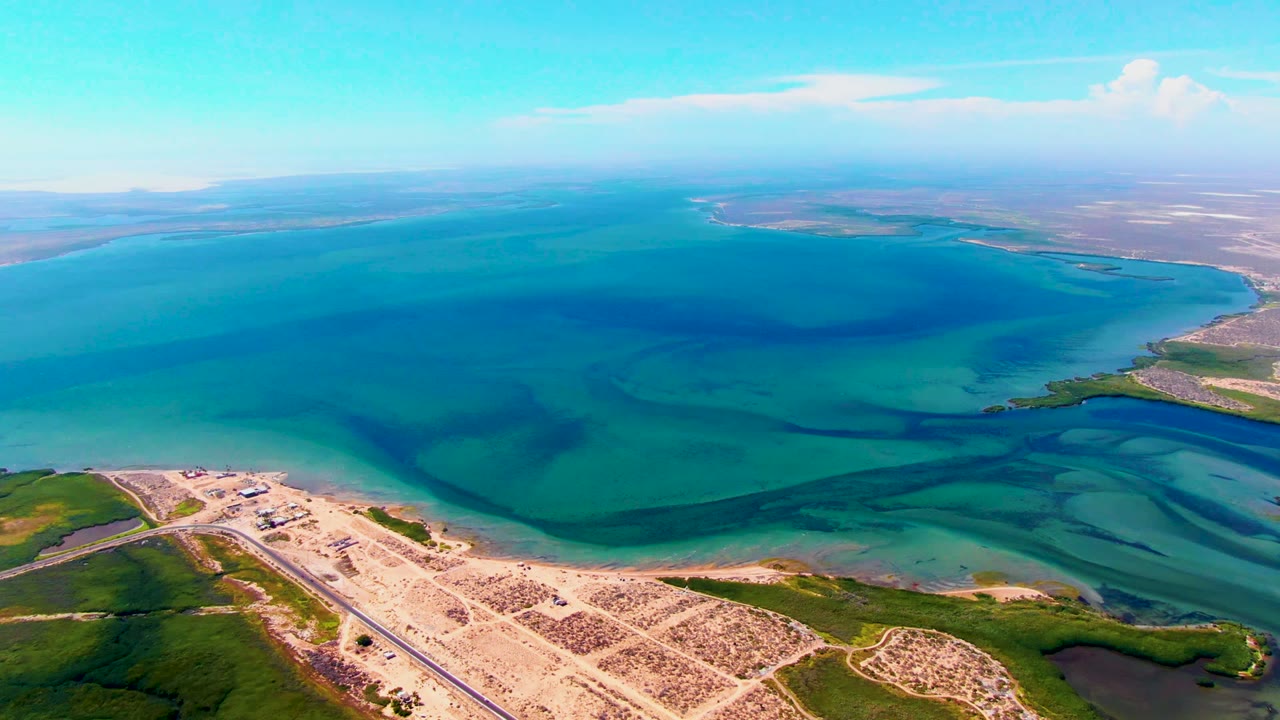 Opening aerial frame over San Carlos, Baja California Sur, with a straight coastal road, pale sand, green shoreline, turquoise water, distant coast, and no visible people.