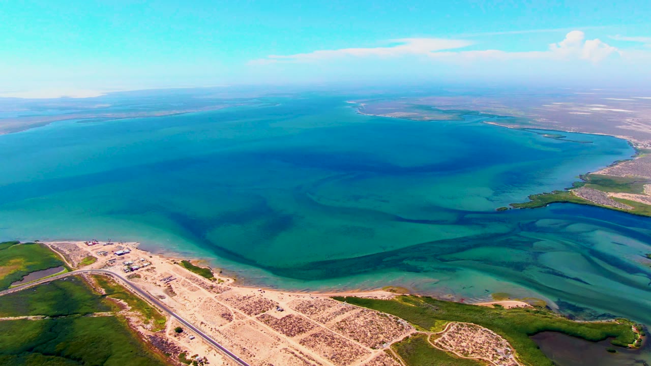 Aerial frame in San Carlos, Baja California Sur, looking across a broad turquoise bay with sandy road edges in the foreground, darker underwater bands, and no visible people.