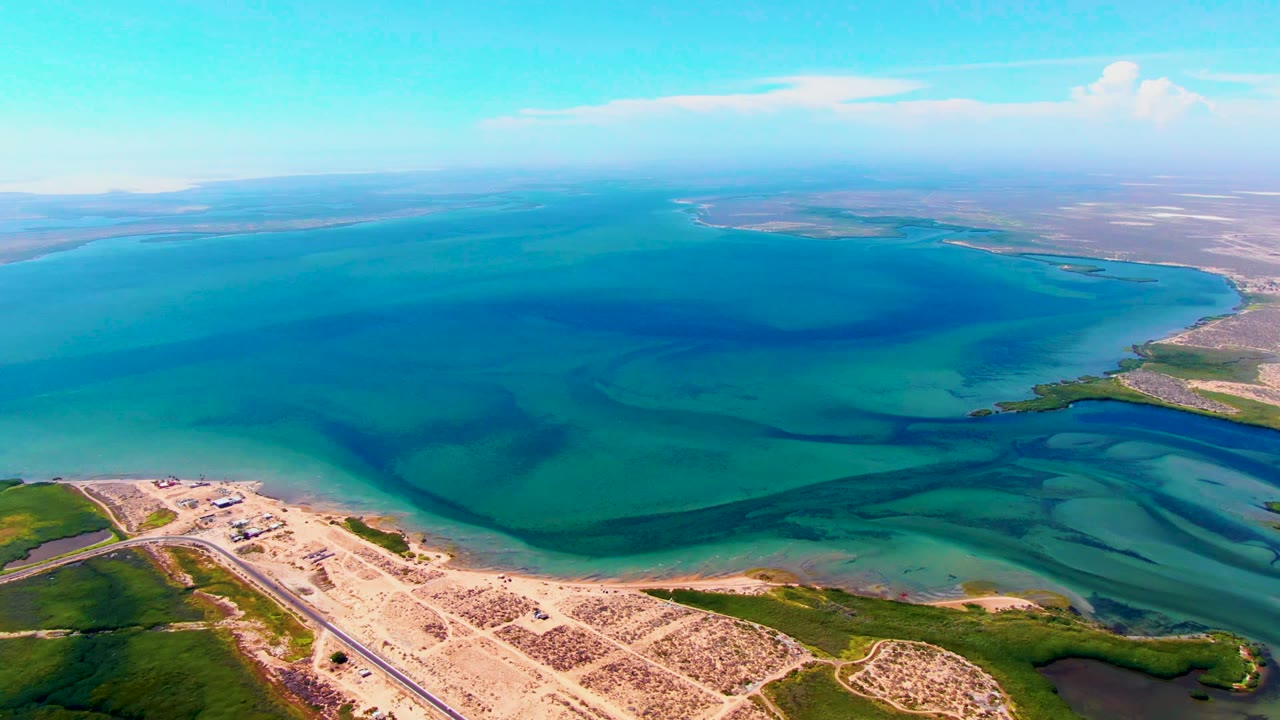 San Carlos aerial frame with the road and sparse sandy development low in frame, green shoreline on the right, layered turquoise water beyond, and no visible people.