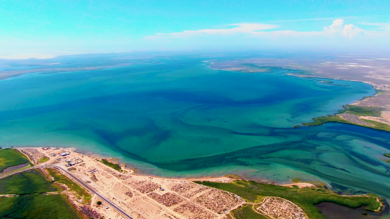 Wide aerial view of clear blue water with sandy shore and road at the bottom.