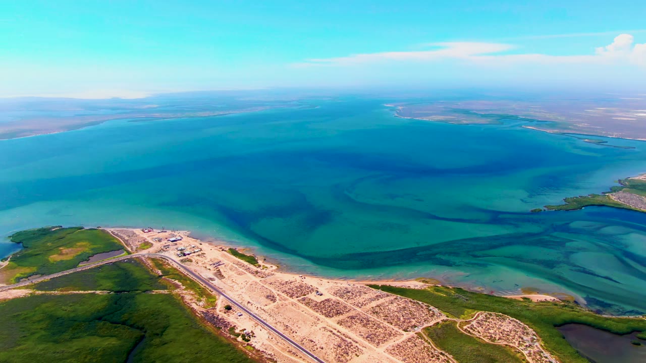 Aerial frame over San Carlos, Baja California Sur showing turquoise ocean, shallow dark water patterns, a sandy developed shoreline, green vegetation, distant low land, and no visible people.