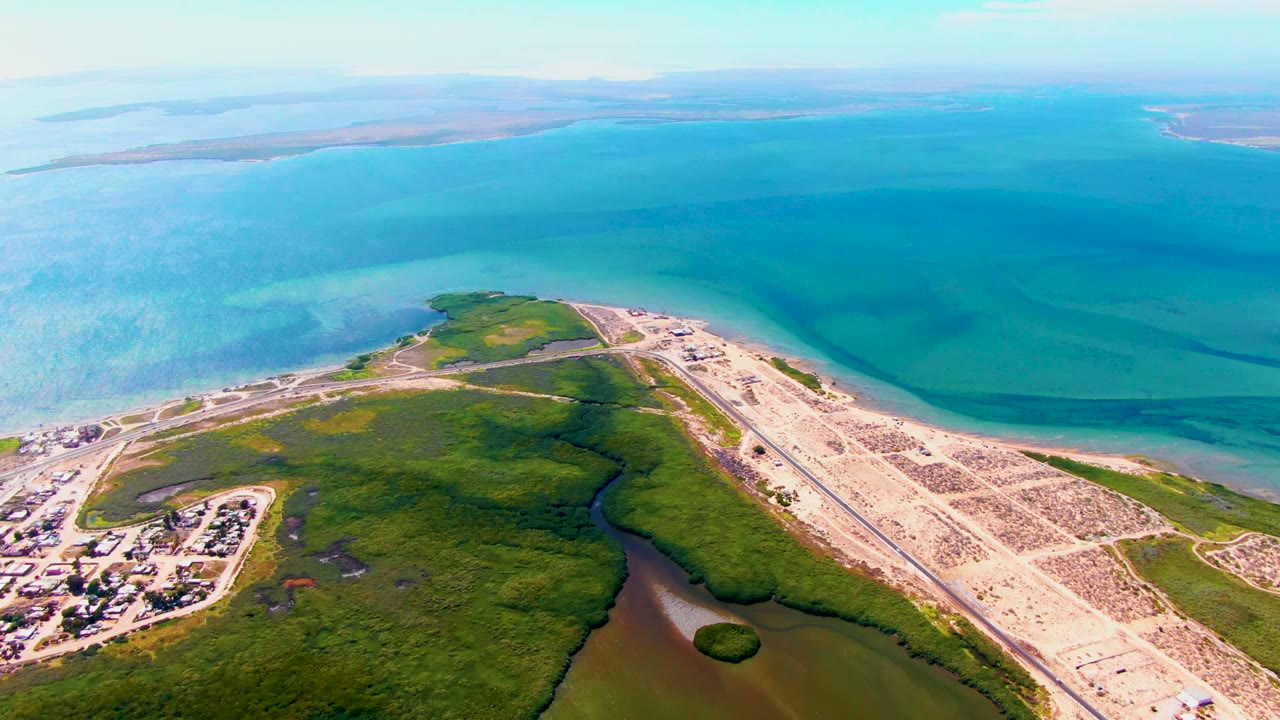 High aerial view over San Carlos showing the runway aligned along the coast, green shoreline vegetation, shallow turquoise water, darker bay patches, and faint islands on the horizon, with no visible people.