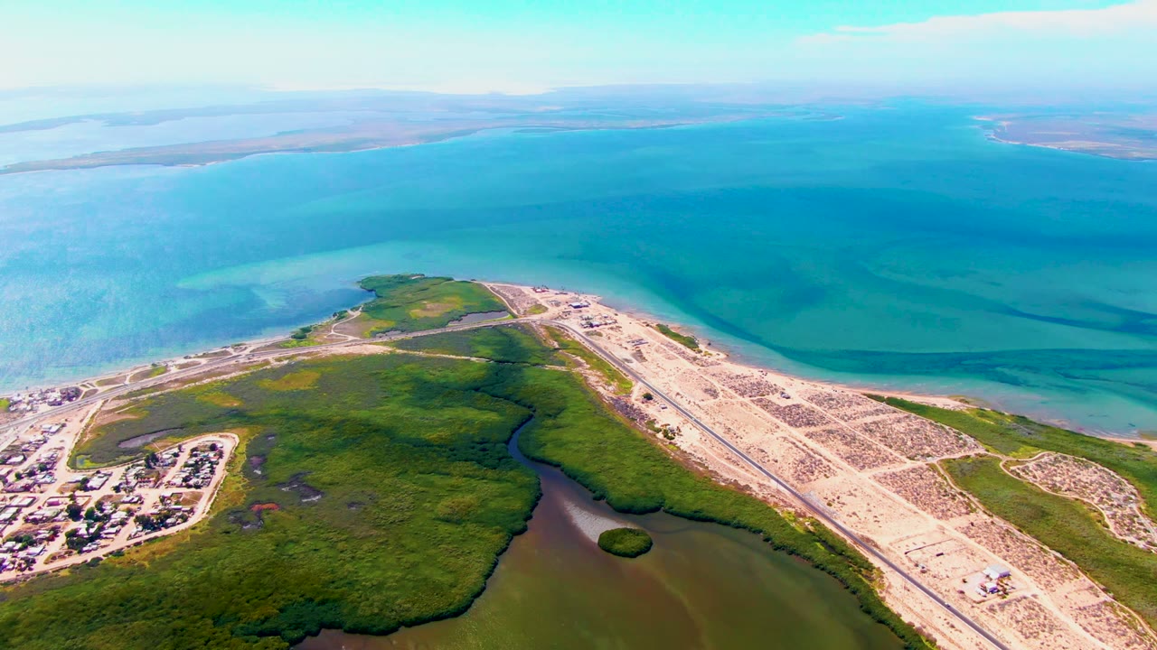 High aerial view over San Carlos, Baja California Sur showing a pale runway construction area beside green wetlands, lagoon water, turquoise ocean, distant land, and no visible people.
