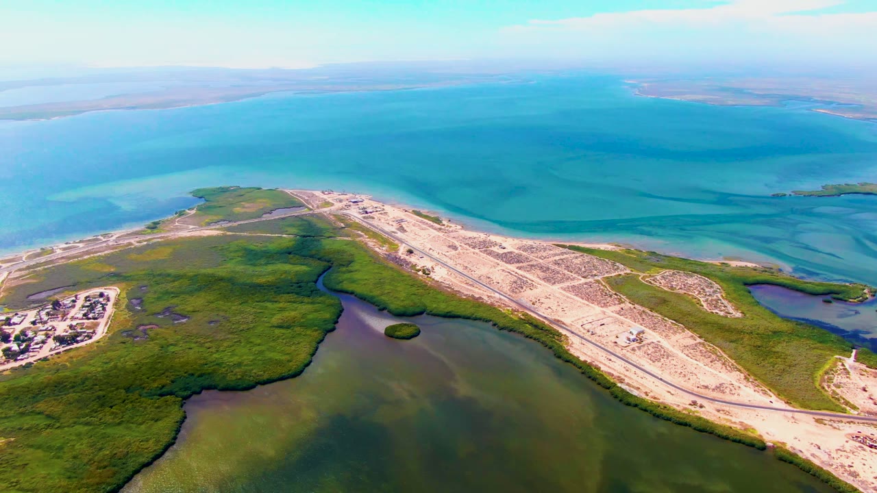 Opening aerial frame over San Carlos, Baja California Sur, with a pale causeway cutting between green marsh, lagoon water, and bright blue ocean, with no visible people.