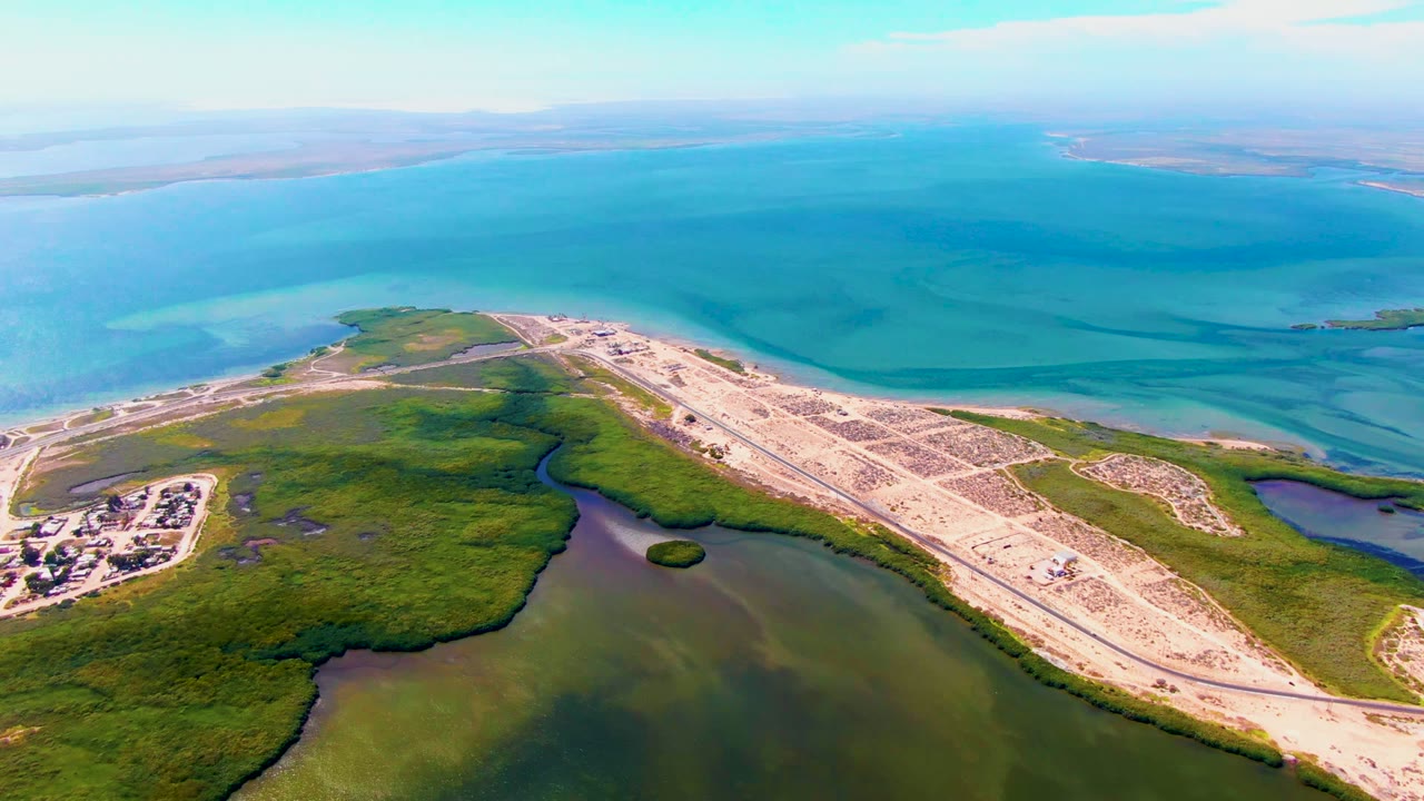 Mid-clip aerial view over San Carlos, Baja California Sur, with the causeway angled across marsh and shallow water toward the broad blue ocean, with no visible people.