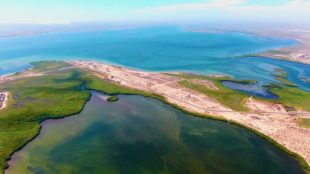 Aerial frame over San Carlos, Baja California Sur, showing dark lagoon water in the foreground, green wetlands, a pale coastal road corridor, turquoise ocean beyond, and no visible people.