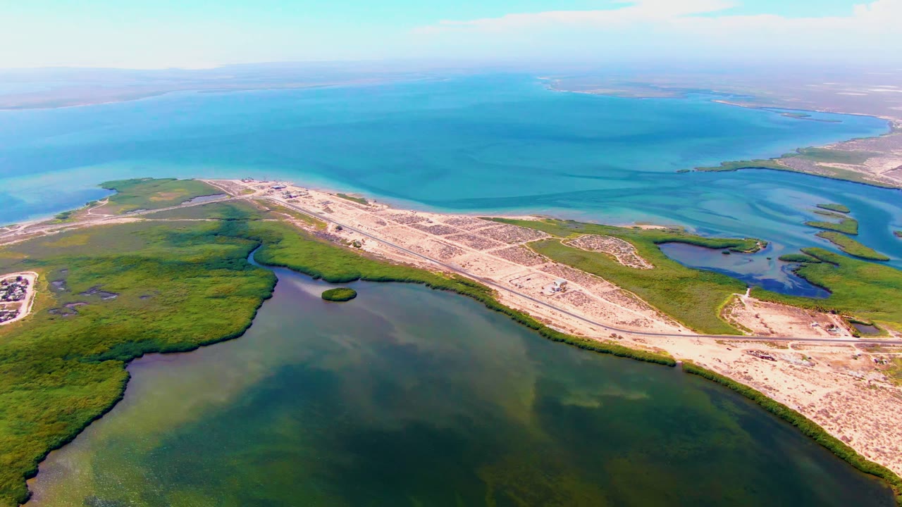 Drone-like aerial frame over San Carlos, Baja California Sur, showing the lagoon foreground, green marsh channels, sandy road alignment, small vehicles, blue ocean, and no visible people.
