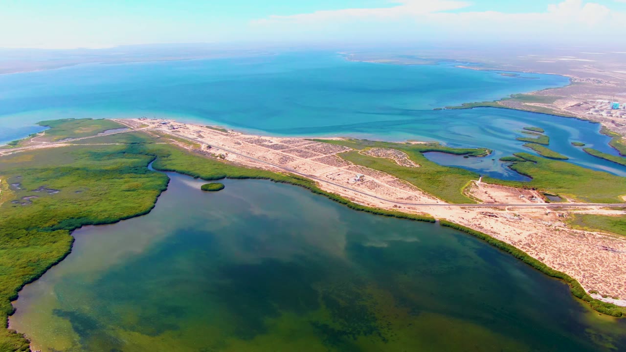 Aerial view over San Carlos, Baja California Sur showing a sandy coastal road between green wetland edges, dark lagoon water, and a bright blue bay, with no visible people.