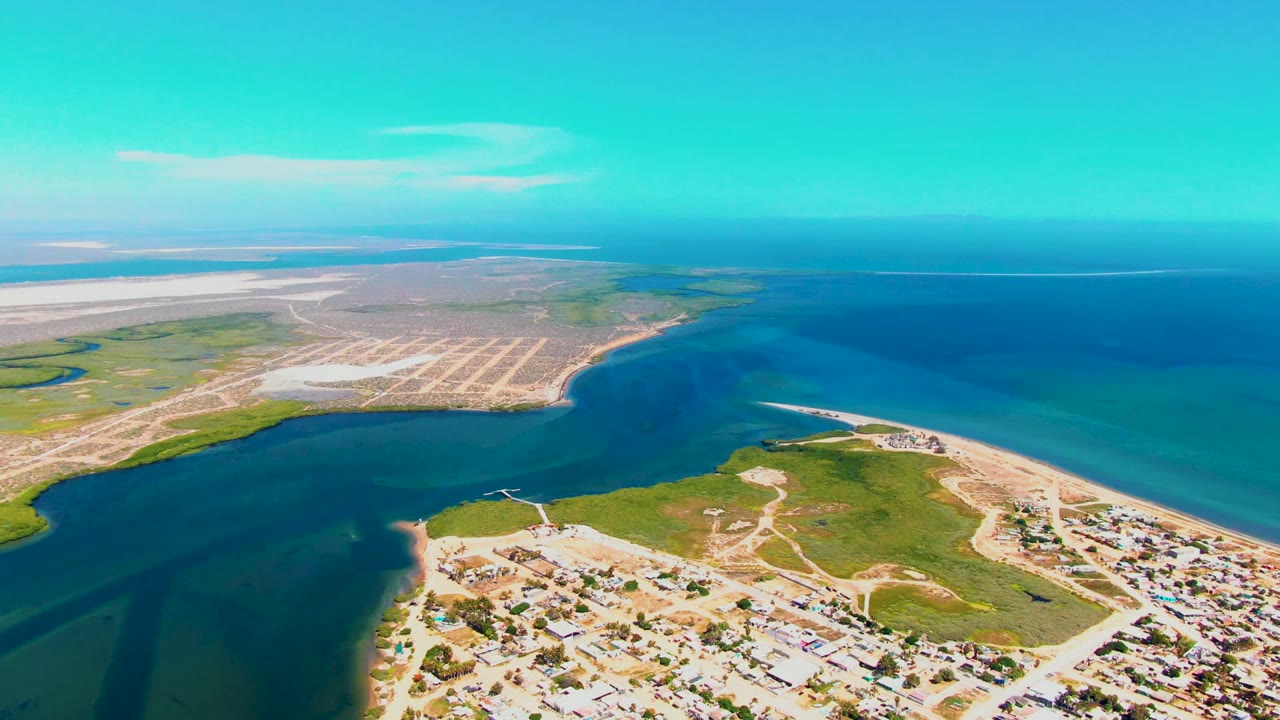 Aerial frame over San Carlos, Baja California Sur showing blue inlet water, pale sand flats, green shoreline vegetation, and small buildings along the lower edge with no visible people.