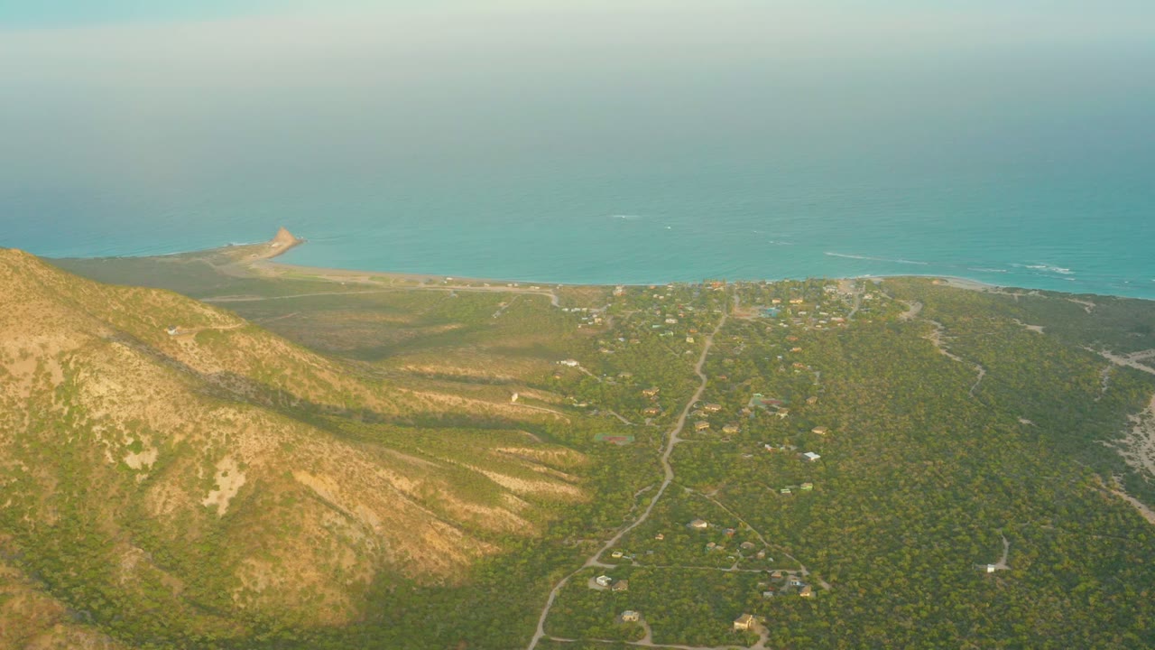 Opening aerial frame over Cabo Pulmo, Baja California Sur, with a tan mountain slope at left, green vegetation and roads below, blue ocean in the background, and no visible people.