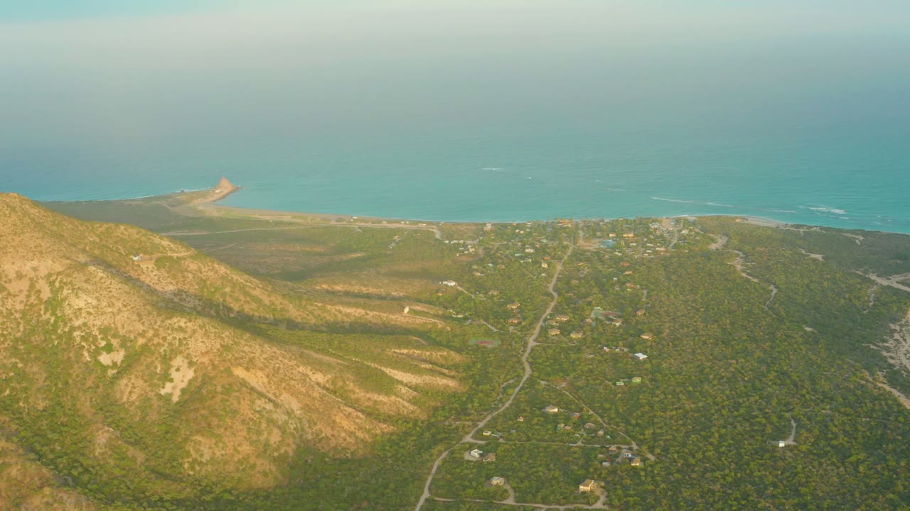 High coastal aerial with hillside, winding roads, small buildings, and ocean water.