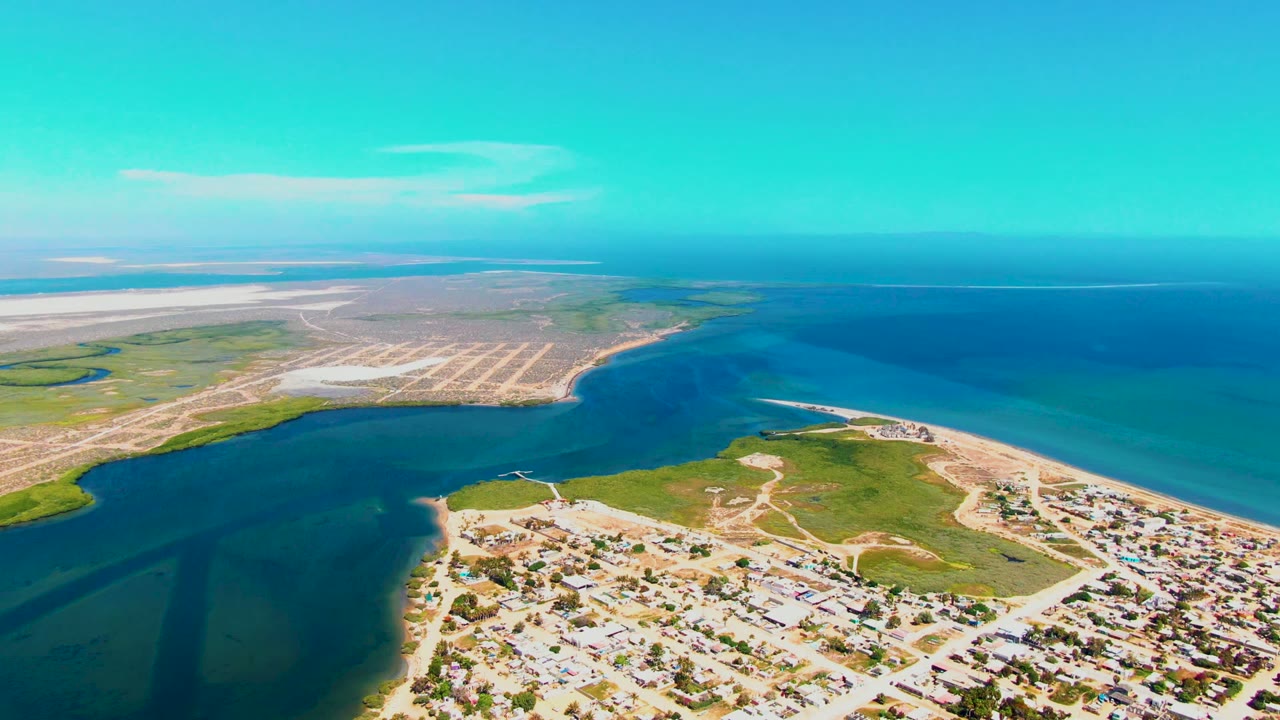 Opening aerial frame over San Carlos, Baja California Sur, with shoreline homes in the foreground, green inlet edges, pale sandy flats, open ocean, and no visible people.