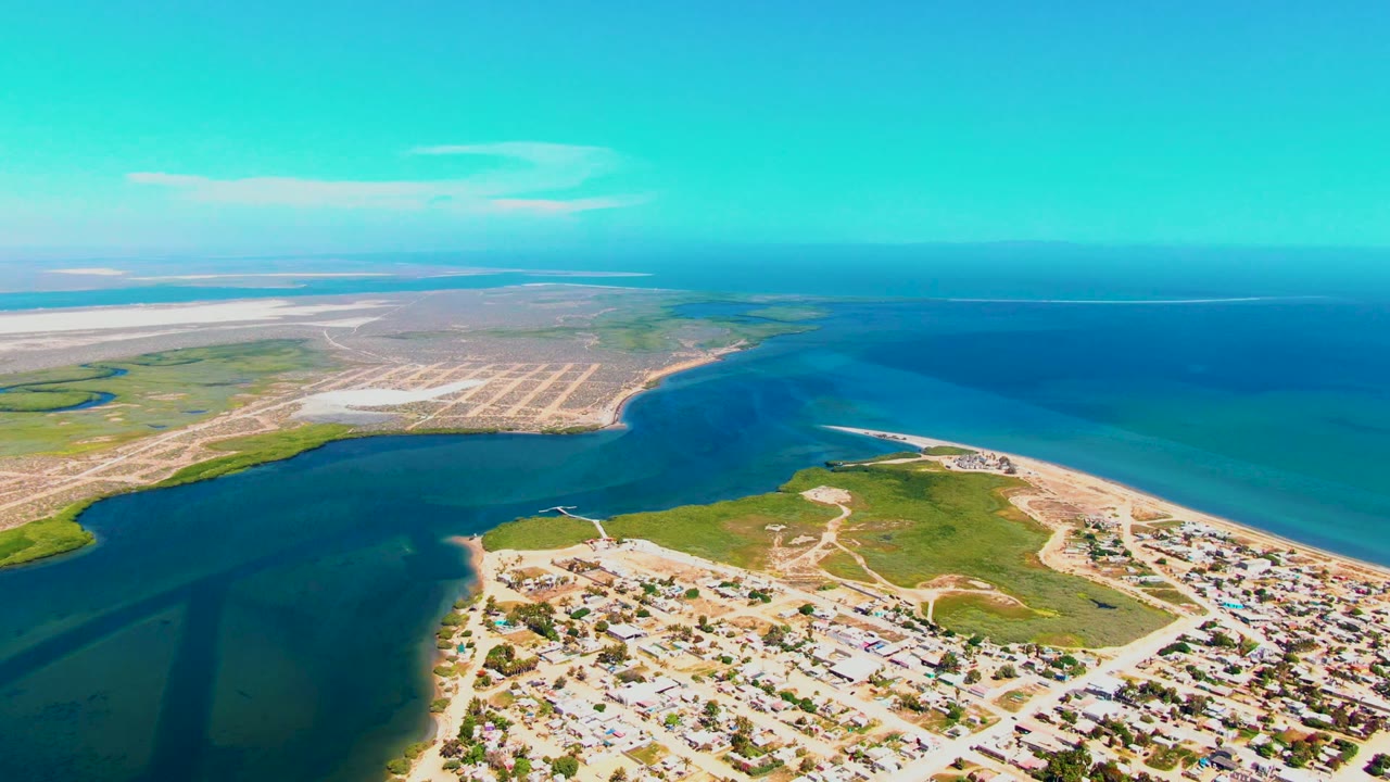 Wide drone view of turquoise inlet water beside sandy shore and houses.