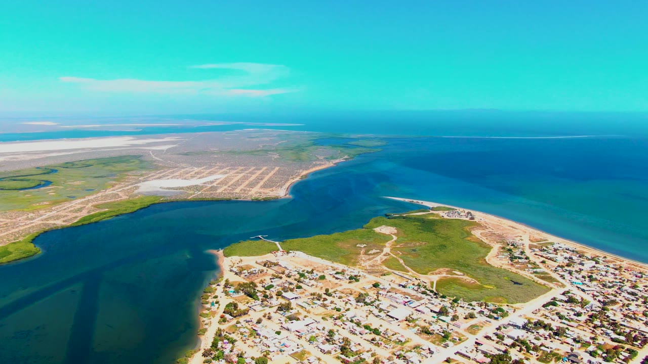 Mid-clip aerial view of San Carlos, Baja California Sur with the inlet cutting through green shore vegetation, houses below, pale sandbars beyond, and no visible people.