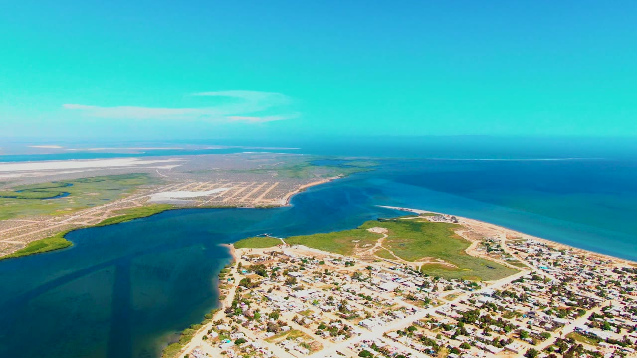 Wide aerial frame over San Carlos, Baja California Sur showing a residential shoreline beside a blue inlet, exposed sand, open ocean, clear sky, and no visible people.