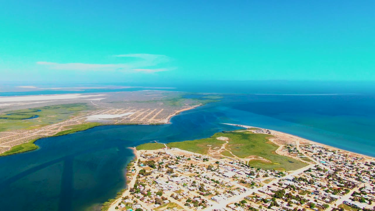 High aerial frame in San Carlos with more open water filling the right side, a sandy shoreline and residential blocks below, green coastal land, clear sky, and no visible people.