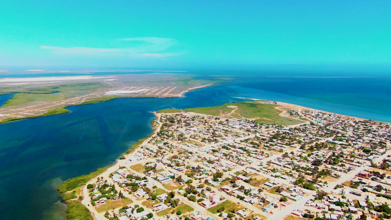 Aerial view over San Carlos, Baja California Sur, with residential blocks in the lower frame, a blue inlet on the left, open ocean on the right, distant land across the horizon, and no visible people.
