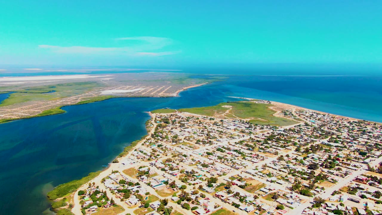 Aerial frame over San Carlos with compact houses and roads at lower right, dark blue inlet water at left, sandy land across the water, open ocean at upper right, and no visible people.