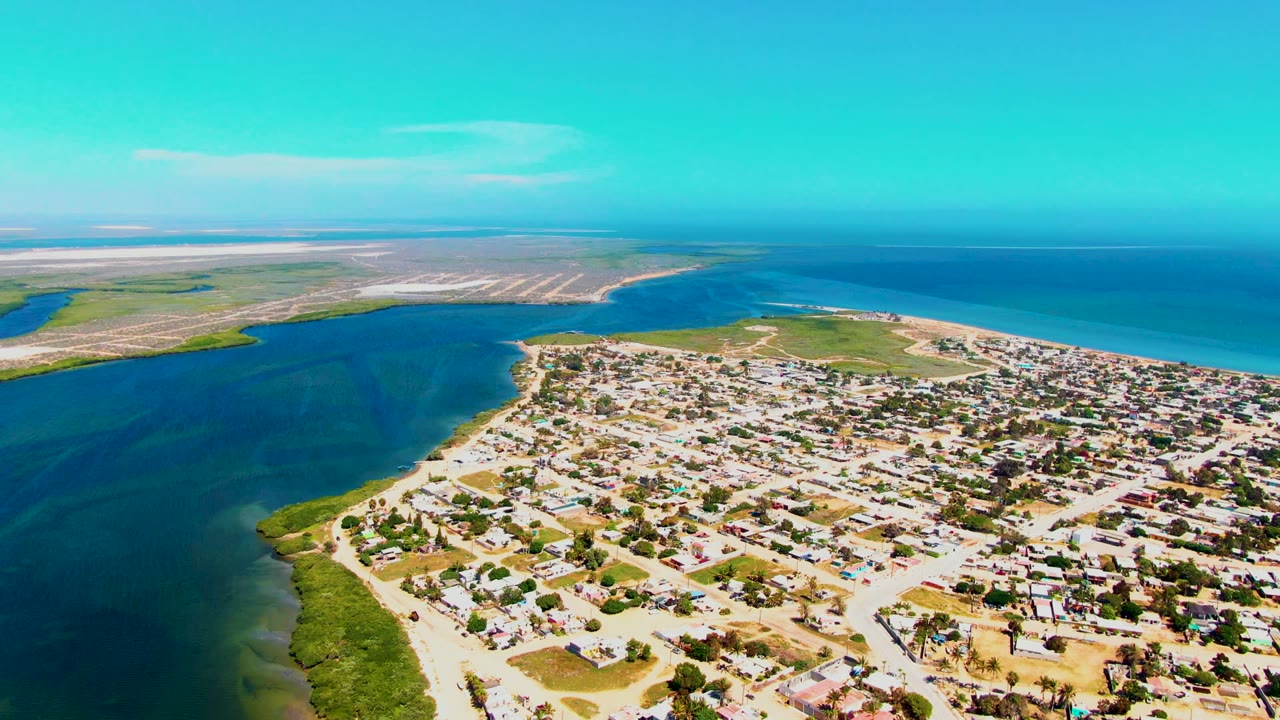 Opening aerial frame over San Carlos, Baja California Sur, with low-rise homes and sandy roads in front of a dark inlet, green shoreline, bright ocean, and no visible people.