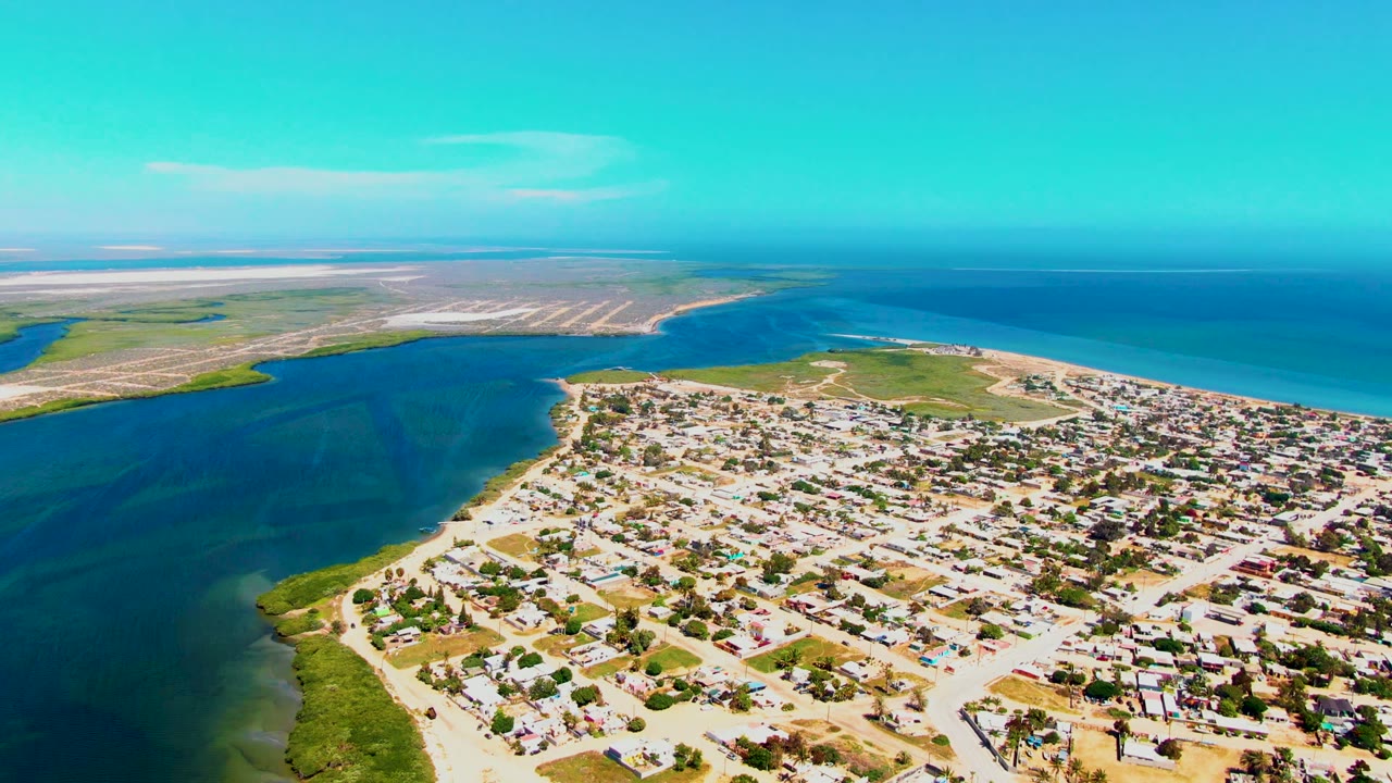 High aerial frame over San Carlos, Baja California Sur, with houses filling the lower right, dark inlet water to the left, a sandy spit near the ocean, and no visible people.