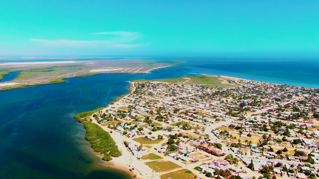 Aerial view over San Carlos, Baja California Sur, showing a blue-green inlet at left, low town buildings at lower right, distant flats, open ocean, and no visible people.