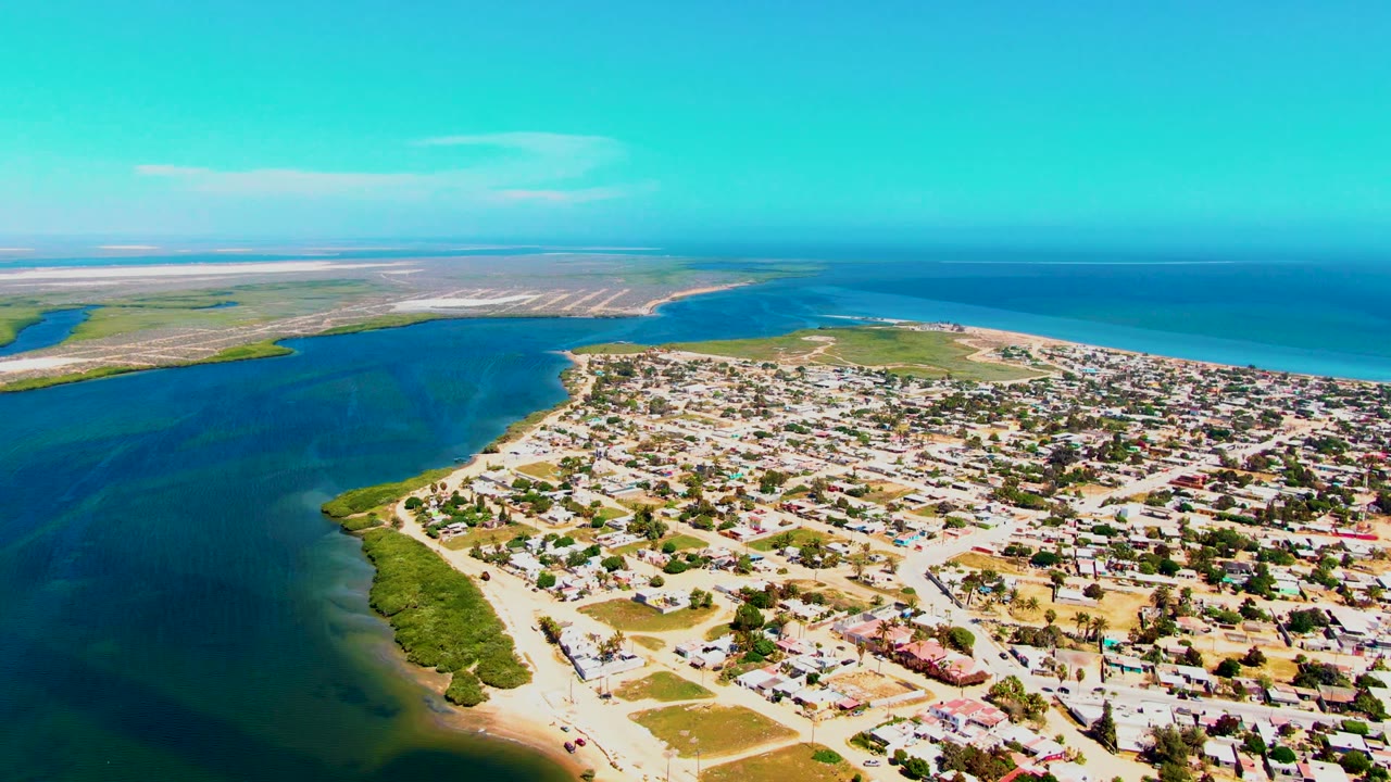 Elevated view of San Carlos with dark water on the left, sandy shoreline and vegetation below, dense low buildings to the right, distant landforms, ocean, and no visible people.