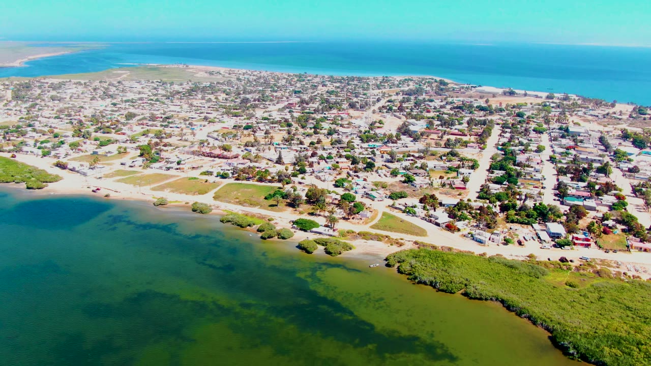 Opening aerial frame over San Carlos, Baja California Sur showing green lagoon water in the foreground, a residential shoreline town, blue ocean beyond, and no visible people.