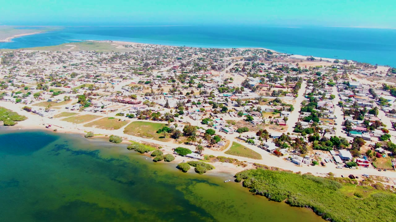 Mid-clip aerial view of San Carlos, Baja California Sur showing lagoon water and vegetation in front, low houses and streets through the center, ocean in back, and no visible people.