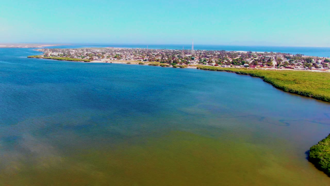 Aerial frame over San Carlos, Baja California Sur, with blue-green water in the foreground, a vegetated shoreline at right, town buildings across the far shore, and no visible people.