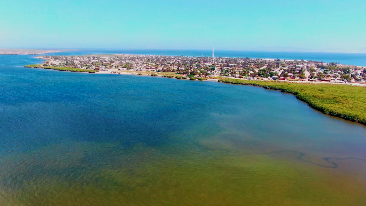 High aerial frame of San Carlos, Baja California Sur, showing darker blue water at left, green shallows near the lower edge, a narrow shoreline, clustered buildings, and no visible people.