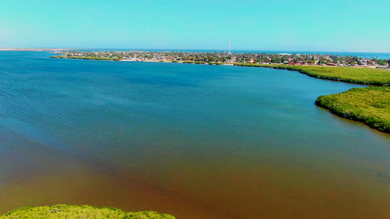 Aerial frame over San Carlos, Baja California Sur showing blue-green estuary water, a green marsh point at right, a distant town and tower on the horizon, and no visible people.