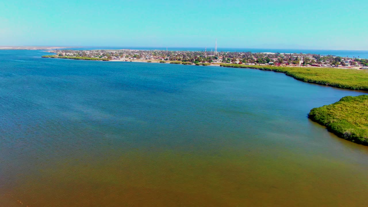 Drone view over San Carlos, Baja California Sur with open blue-green water in the foreground, a curved marsh shoreline on the right, a distant coastal town, a tall tower, and no visible people.