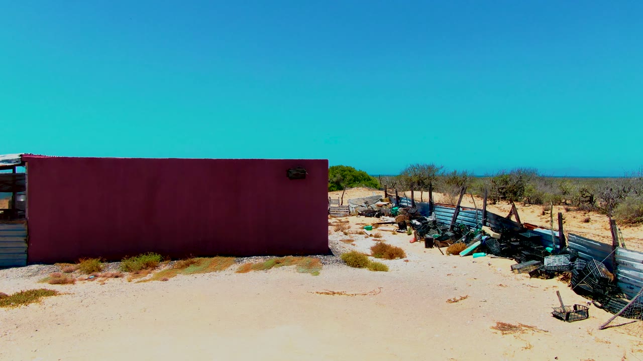 Wide view in San Carlos, Baja California Sur of a maroon container, sandy foreground, fencing, scrap debris, scrub, and no visible people under a clear blue sky.