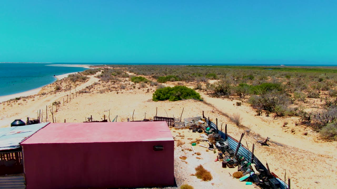 Wide elevated view in San Carlos, Baja California Sur with a red beach structure low in frame, sandy paths, fence posts, dry vegetation, ocean at left, and no visible people.