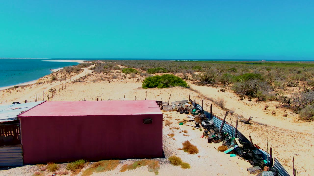 Red structure on sandy beach with fence, netting, scrub, and ocean behind.