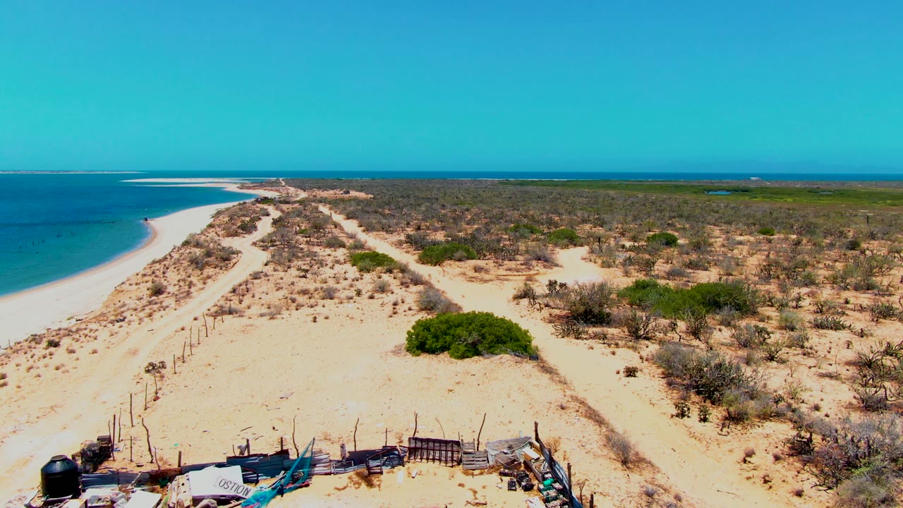 Drone view of beach dunes, scrubland, and a partial structure at the bottom edge.