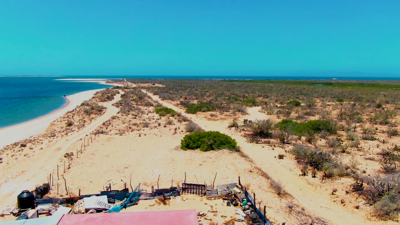 San Carlos aerial frame with turquoise water at left, sand dunes crossing the middle, desert scrub across the right side, fencing and materials near the bottom, and no visible people.