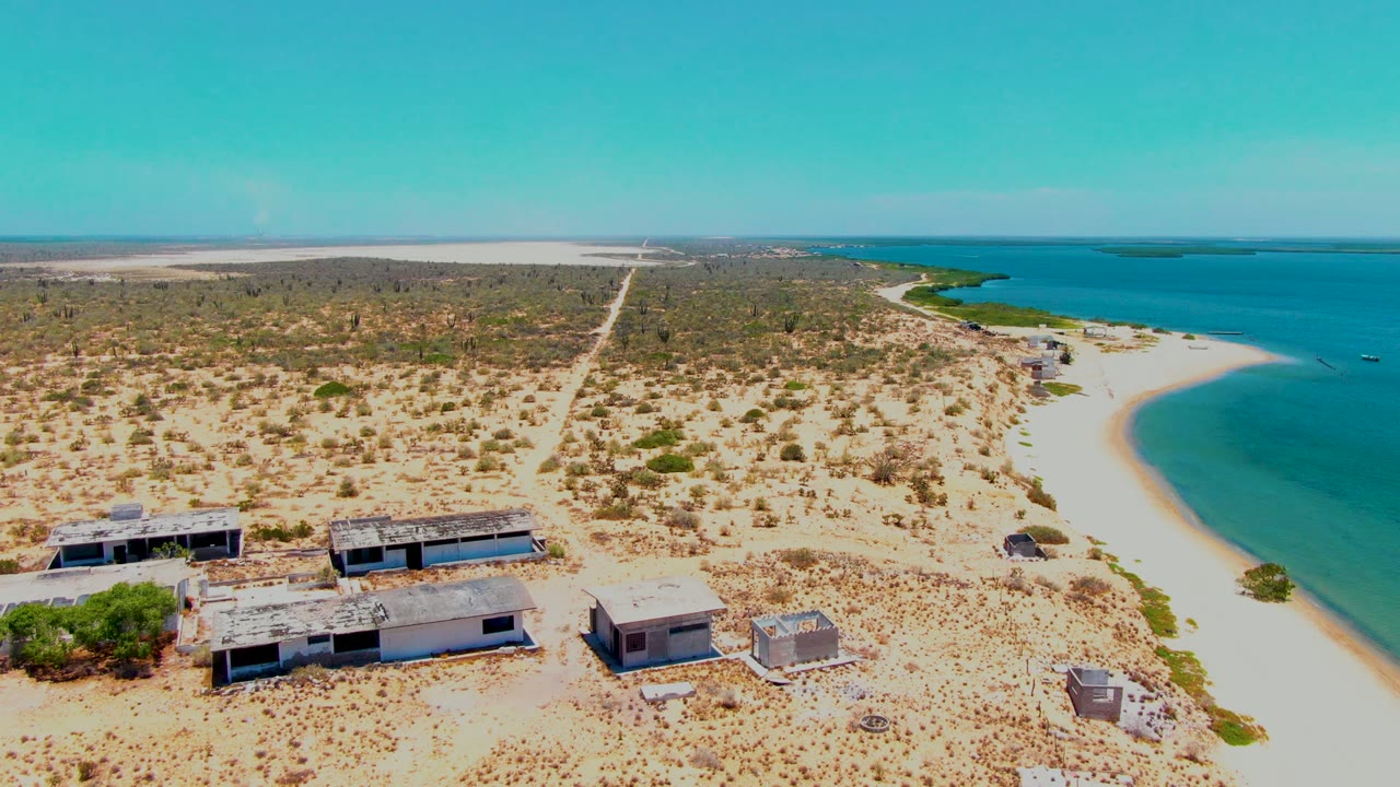 Aerial frame over San Carlos, Baja California Sur showing abandoned flat-roofed structures in sandy scrub beside a curved beach and blue ocean, with no visible people.