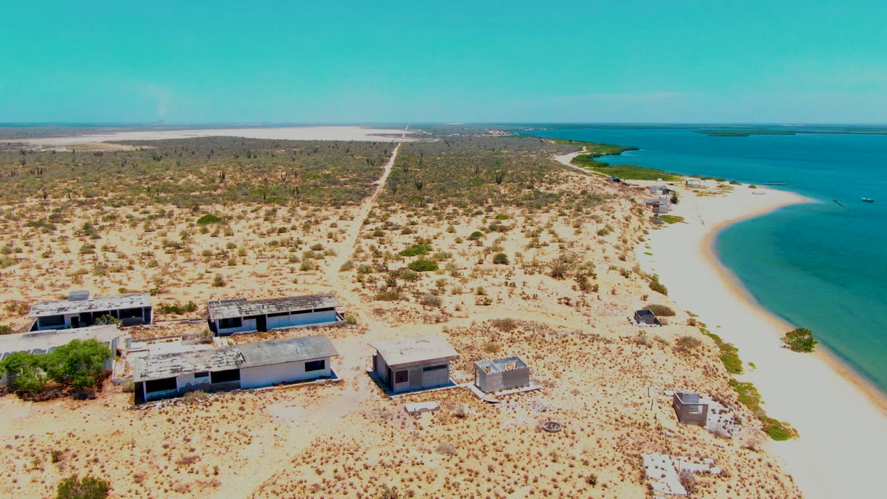 High aerial frame over San Carlos, Baja California Sur with low abandoned buildings near the lower edge, sparse desert brush across the center, and blue water along the right, with no visible people.