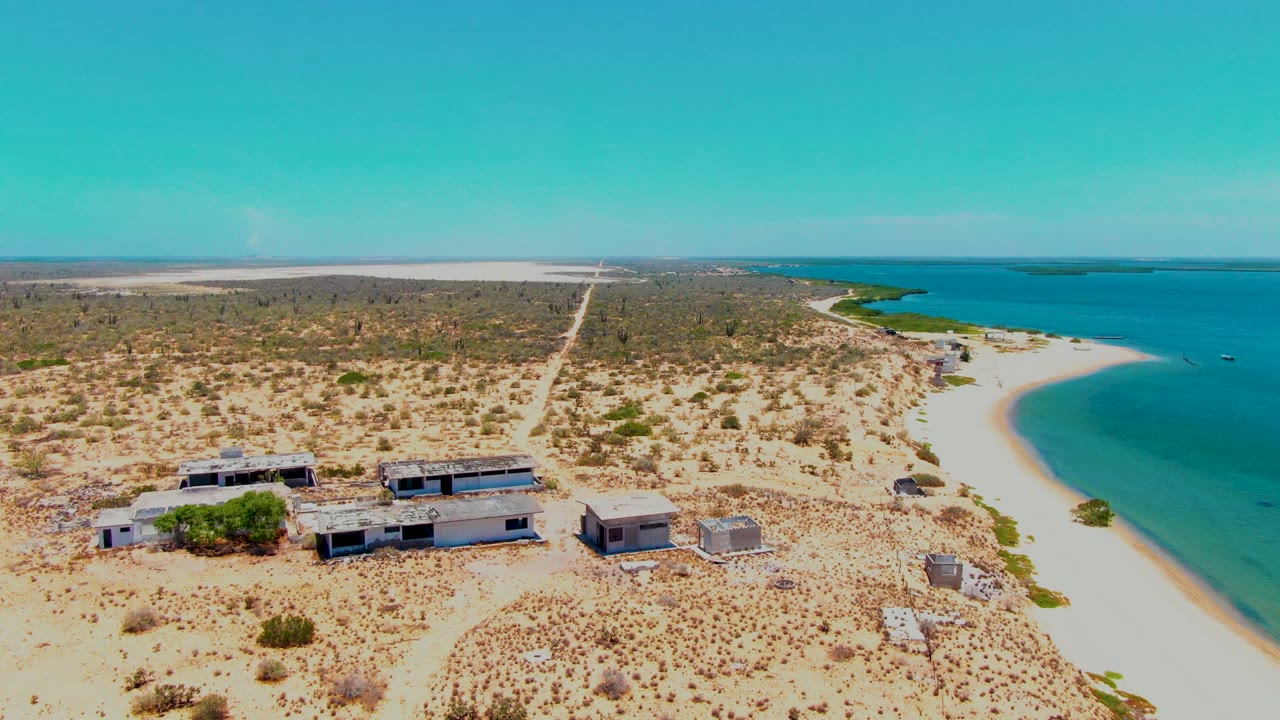 Aerial frame over San Carlos, Baja California Sur showing low rectangular coastal buildings on pale arid land beside a curved sandy beach and turquoise water, with no visible people.