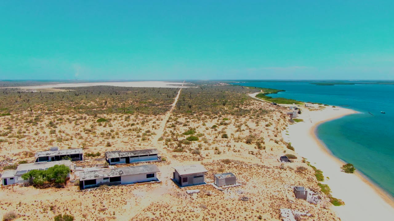 Drone-like frame over San Carlos, Baja California Sur with several low buildings on sandy ground, a curved white beach to the right, green-blue water beyond it, and open scrubland stretching inland, with no visible people.