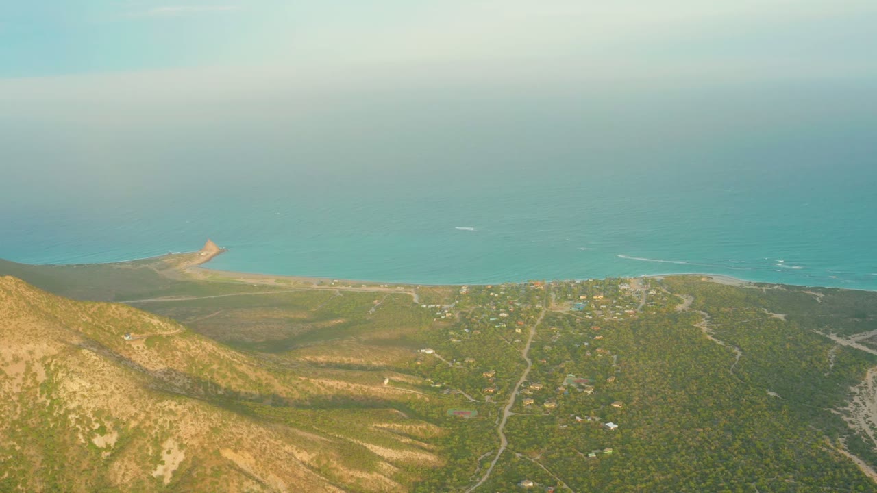 Opening aerial frame over Cabo Pulmo, Baja California Sur, with dry hills in the foreground, green scrubland near the shore, blue ocean beyond, and no visible people.
