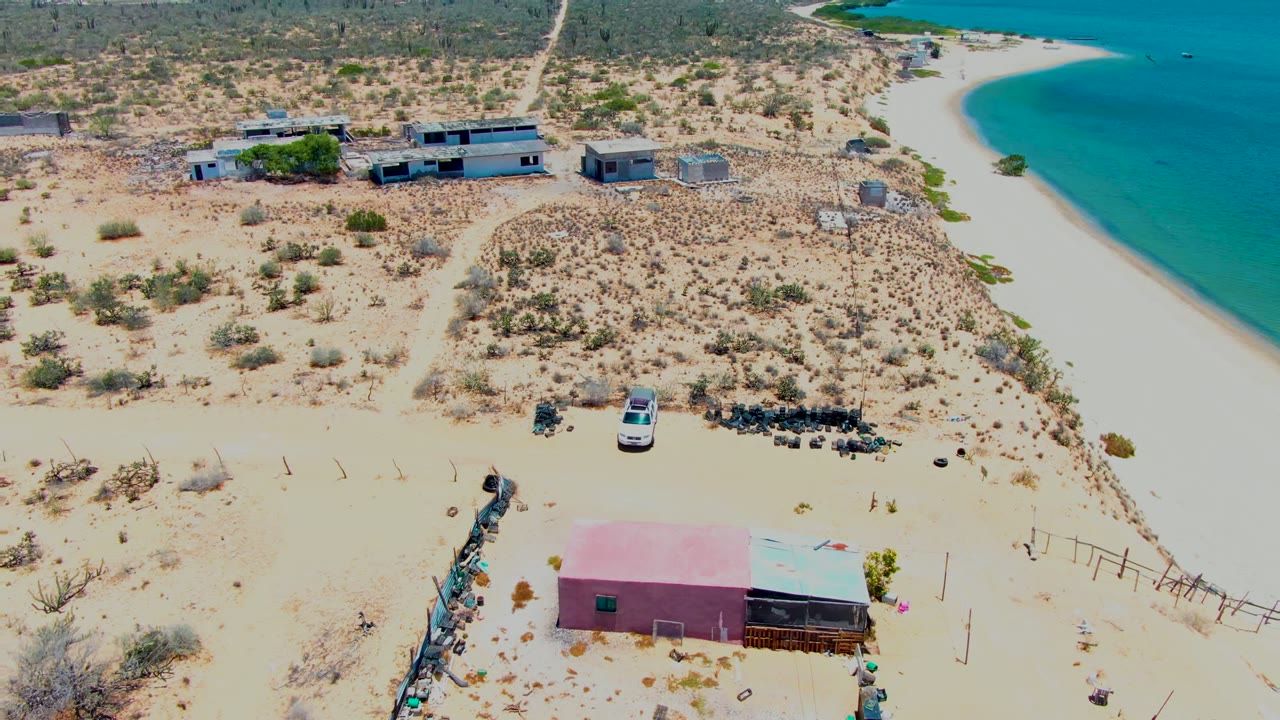 Aerial frame over San Carlos, Baja California Sur showing sandy scrubland, low buildings, a pink-roofed structure near the bottom edge, a white vehicle, a curving beach, turquoise water, and no visible people.