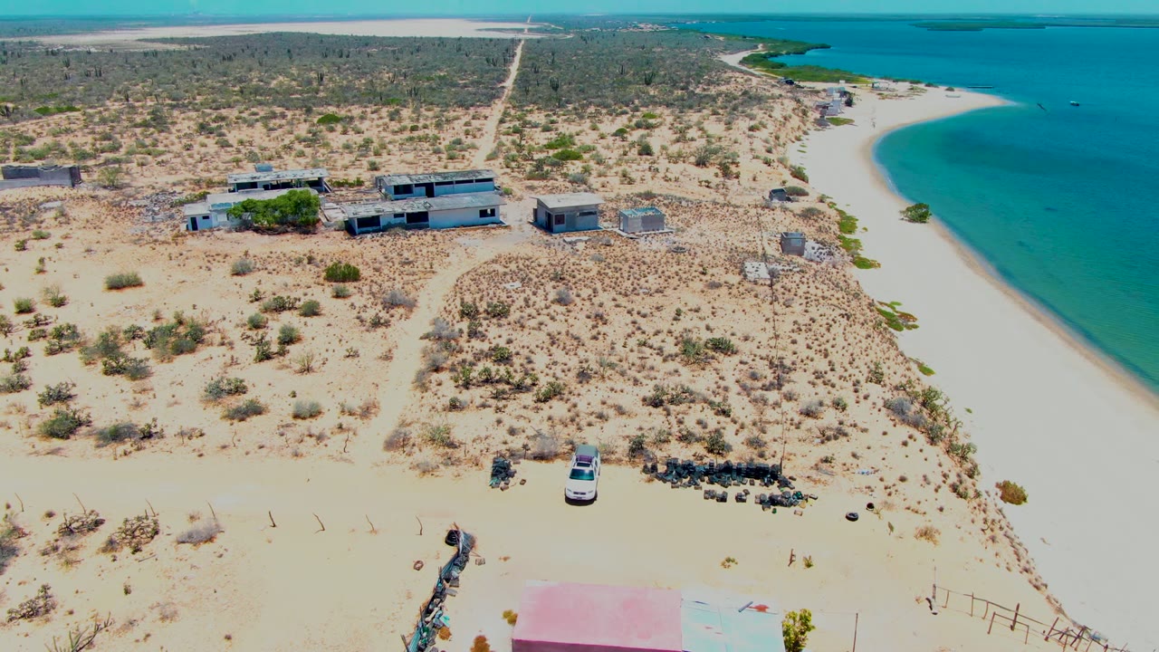 Higher aerial frame in San Carlos, Baja California Sur showing the pink-roofed structure partly cropped at the bottom, clustered low buildings, scrub-covered sand, a straight road running inland, turquoise water, and no visible people.