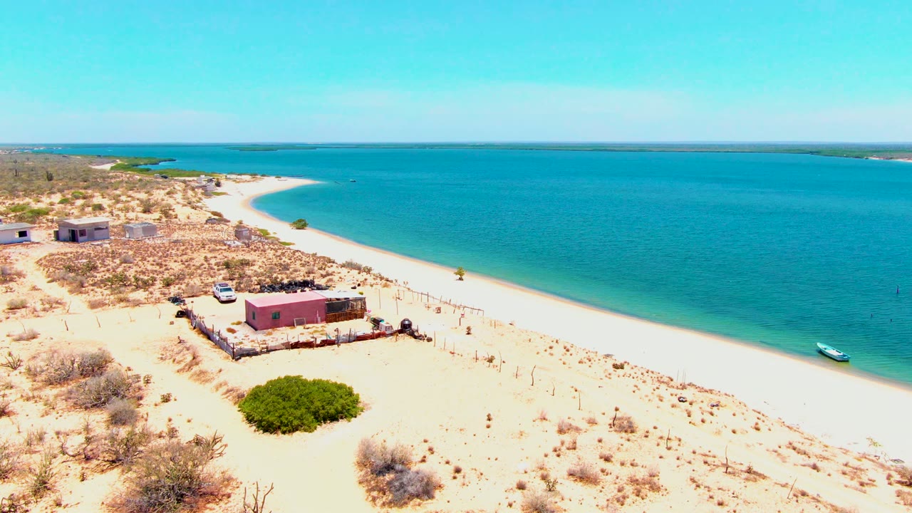 Elevated beach view with a pink shore building, white car, green bush, and small boat near calm water.