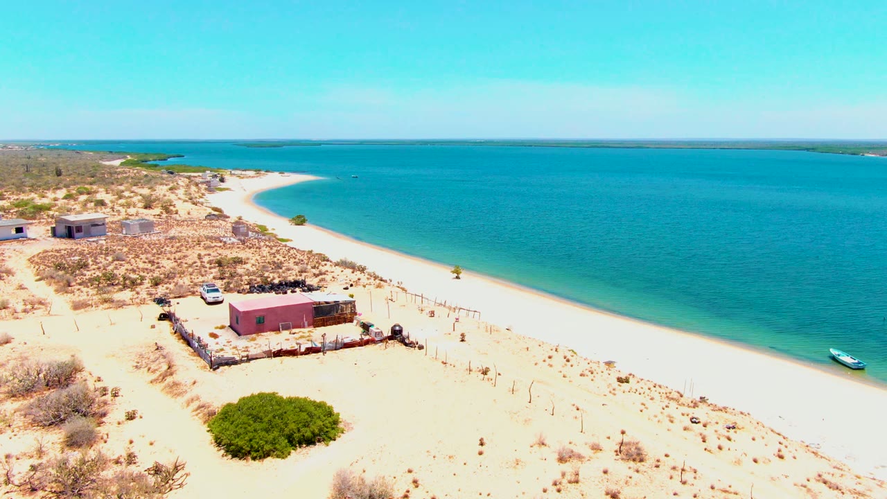 Aerial view in San Carlos with the pink building and fence line below center, the white car set back on sand, sparse desert vegetation, turquoise water, and no visible people.
