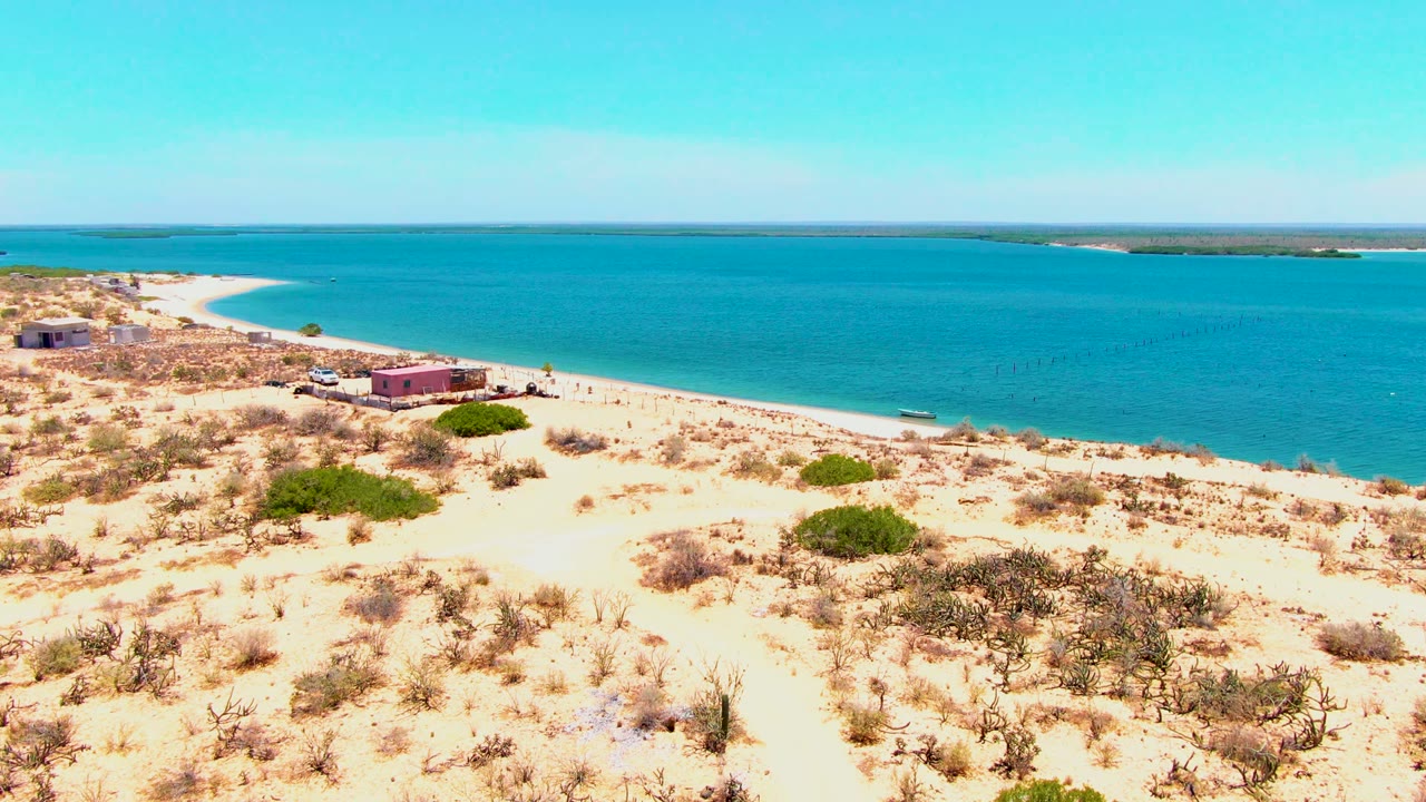 Aerial view over San Carlos, Baja California Sur, with pale sand and scattered shrubs in the foreground, small beachside buildings near the shore, a white vehicle by the structures, blue water beyond, and tiny people visible on the beach.