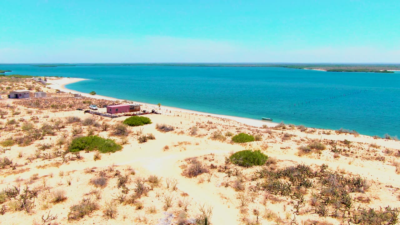 Elevated shoreline view with sand, shrubs, beach buildings, turquoise water, and a small boat.